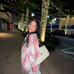 Cristina Hernandez smiles while standing on a decorated street at night. She is wearing a pink and white plaid shirt and carries a white handbag. The street is lined with trees wrapped in lights, and buildings are visible in the background.